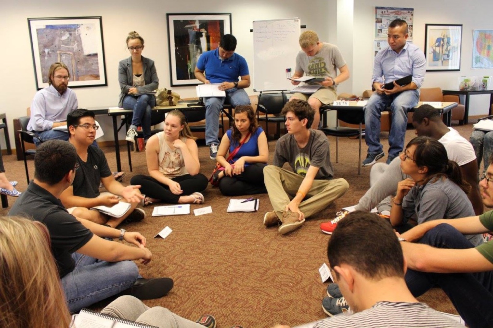 A wide view of a discussion circle, participants seated on the floor and around the edge of the room.